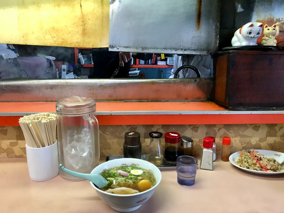 카운터 자리에 앉아 본 모습 — 앞에 놓인 라멘 한 그릇과 나무젓가락 통, 물병, 양념병, 너머로 김이 오르는 주방